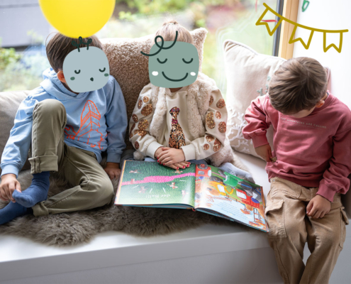 Ein Mädchen und zwei Jungs sitzen auf einer Bank mit einem Buch über ihnen hängen Luftballons