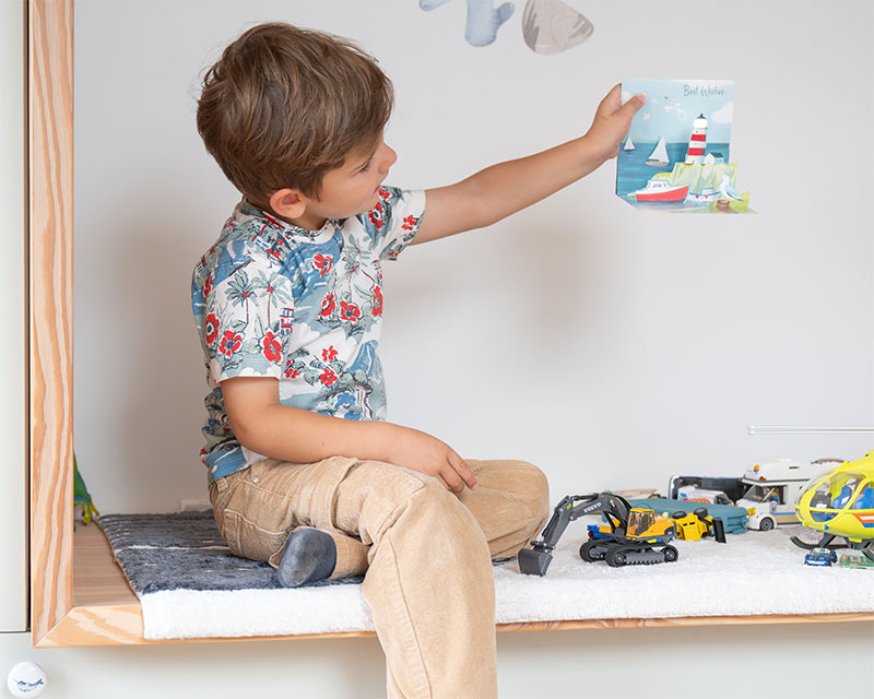 Ein Junge sitzt in einer Nische, vor ihm viele Spielsachen und er hält eine 3D-Karte von einem Leuchtturm hoch