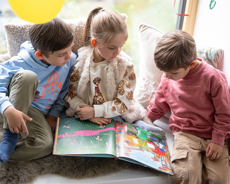 Ein Mädchen und zwei Jungs sitzen auf einer Bank mit einem Buch über ihnen hängen Luftballons
